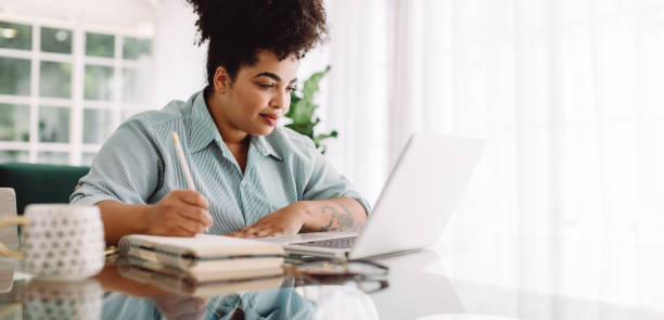 Businesswoman looking busy working from home Business woman working from home writing notes while looking at laptop. Confident woman sitting at desk using laptop and taking notes. online education stock pictures, royalty-free photos & images