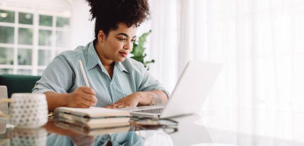 Businesswoman looking busy working from home Business woman working from home writing notes while looking at laptop. Confident woman sitting at desk using laptop and taking notes. writing stock pictures, royalty-free photos & images