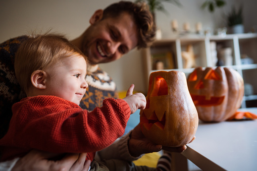 Handsome young Caucasian father having fun while preparing a pumpkin with his baby son for his first Halloween holiday.