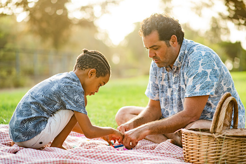 Adult man and black boy playing with toys while sitting on checkered blanket near basket during picnic in summer in park