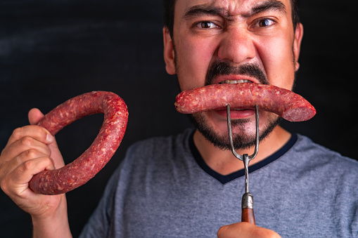 A bearded, hungry man greedily eats a huge piece of sausage and holds a sausage ring in his other hand.