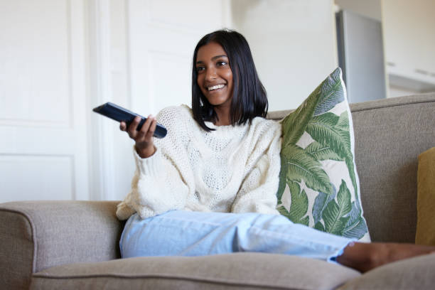 shot of a beautiful young woman holding a remote control while sitting on the couch at home - controlo remoto imagens e fotografias de stock