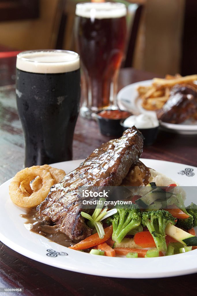 Delicious steak served with vegetables, onion rings and beer 10 oz. New York Striploin Angus steak topped with Irish whiskey gravy, sautéed mushrooms and onion rings. Served with fresh vegetables and mashed potatoes. Goes best with Guinness. Focus on the front of the steak. Steak Stock Photo