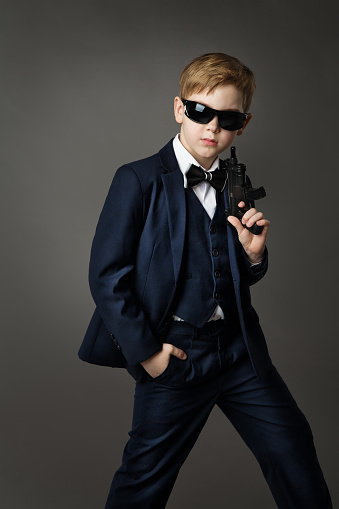 Little Kid Boy In Black Suit and Sunglasses holding Shooting Gun. Elegant Fashion Child in Tuxedo and Bowtie over Gray