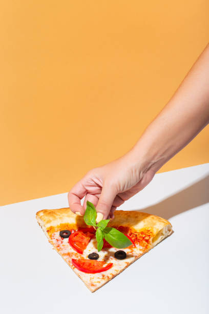Woman hand putting basil leave on slice of pizza. Homemade Italian style pizza with olives, tomatoes and basil stock photo