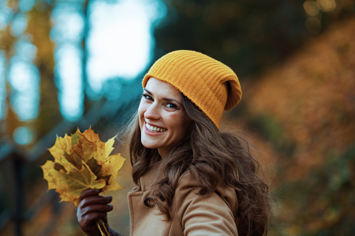 Hello november. happy 40 years old woman in beige coat and orange hat with autumn yellow leaves outside in the city park in autumn.