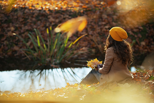 Hello september. Seen from behind stylish woman in beige coat and orange hat with autumn yellow leaves sitting outdoors in the city park in autumn.