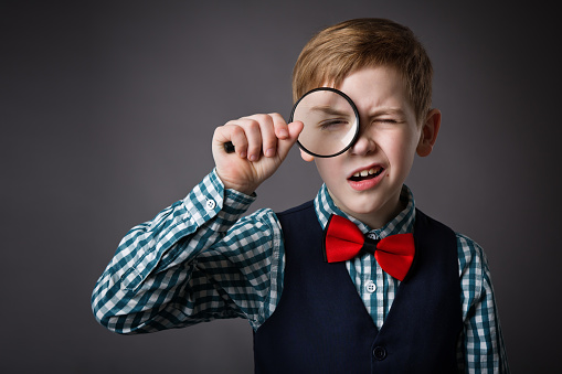 Little Kid Looking Through Magnifying Glass Lens. Funny Surprised Child Holding Loupe. School Boy in Smart Casual Clothing over Gray Studio Background