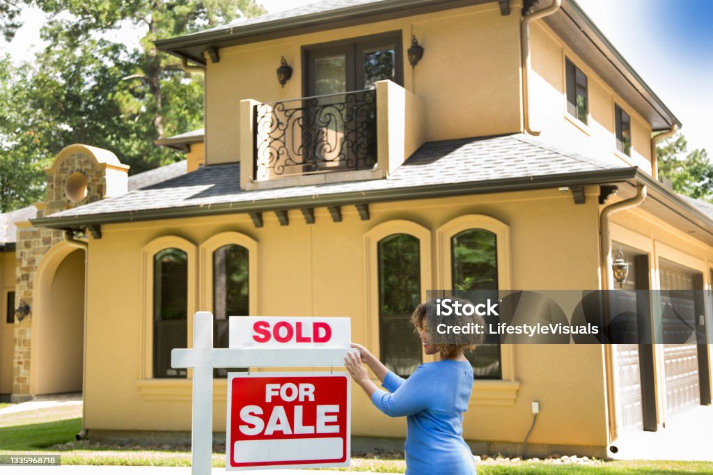Lovely young adult Real Estate Agent standing beside her for sale sign in front yard of home.  She adds the SOLD sign and wears a blue top and jeans. For Sale Sign Stock Photo