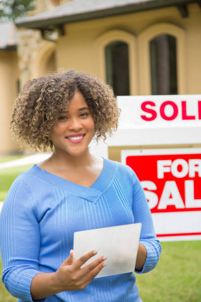 Lovely young adult Real Estate Agent standing beside her for sale sign in front yard of home.  She holds a new translucent digital tablet and wears a blue top and jeans.   She smiles as she looks at the camera. Lovely young adult Real Estate Agent standing beside her for sale sign in front yard of home.  She holds a new translucent digital tablet and wears a blue top and jeans.   She smiles as she looks at the camera. real estate outdoors vertical usa stock pictures, royalty-free photos & images