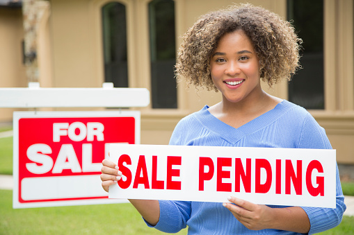 Lovely young adult Real Estate Agent standing beside her for sale sign in front yard of home.  She holds a sale pending sign she plans to place on the top of the sign post.  She wears a blue top and jeans and smiles for the camera.
