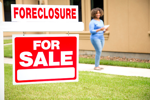 Lovely young adult Real Estate Agent standing in background of her foreclosure sign in front yard of home.    She wears a blue top and jeans and carries a digital tablet.