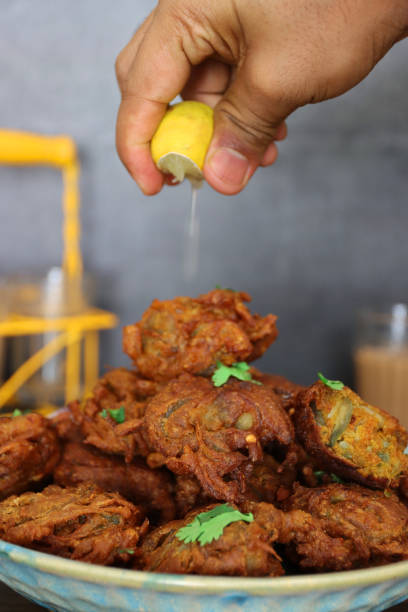 Stock photo showing a close-up view of a batch of homemade crispy onion bahjees in a pile. imagen persona irreconocible exprimiendo jugo de una rodaja de limón sobre una pila de bhajis / pakoras de cebolla crujiente casera adornadas con hojas de cilantro en un plato estampado, bocadillo vegetariano de cocina india, vasos de té chai de fondo, e