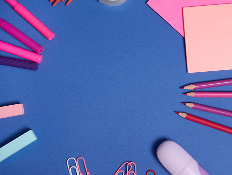 Flat lay composition with stationery or school supplies in pink purple shades scattered in a circle on a blue background with copy space. Teachers day concept