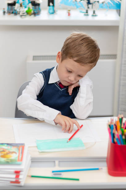 istock A cute boy first grader in a school uniform does homework while sitting at a desk with a pencil in his hands at home. Remote education. Selective focus. Close-up 1335973740