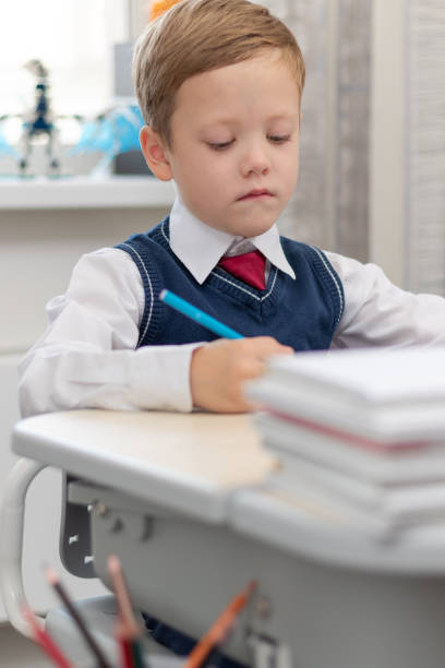 A cute boy first grader in a school uniform does homework while sitting at a desk with a pencil in his hands at home. Remote education. Selective focus. Close-up