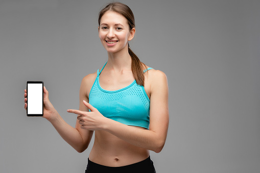 Portrait of nice young woman in sportswear at the studio with gray background. Concept with copy space. She is posing for camera. She have sports athletic body. Girl showing screen on mobile phone