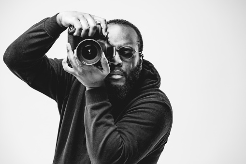 Fotógrafo profesional en el estudio, hombre afroamericano usar sudadera con capucha negra y gafas de sol con cámara digital está trabajando. Fotografía conceptual en blanco y negro photo