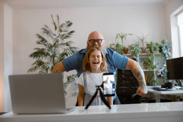 Laughing daughter with funny daddy pose together shooting new video for blog at home Laughing little daughter with funny bearded daddy pose together shooting new video for blog with mobile phone in light room vlogging photos stock pictures, royalty-free photos & images