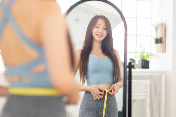 Jeune femme mesurant la taille avec du ruban adhésif debout devant le miroir. Perte de poids. - Photo