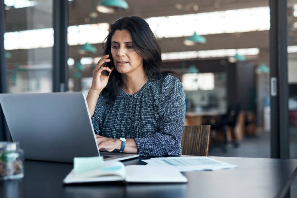 aufnahme einer reifen geschäftsfrau mit laptop und smartphone in einem modernen büro - websurfen stock-fotos und bilder