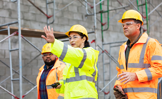 A multi-ethnic group of three workers at a construction site, all looking in the same direction. They are wearing hard hats, safety goggles and reflective clothing. A concrete block wall with scaffolding is out of focus behind them. The main focus is on the Hispanic woman, in her 40s, who is standing in the middle, hand raised in a stop gesture.