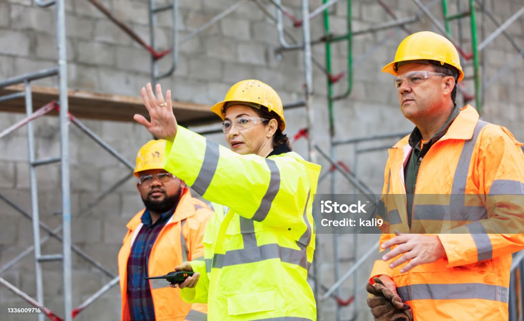 Hispanic woman leads crew at building construction site A multi-ethnic group of three workers at a construction site, all looking in the same direction. They are wearing hard hats, safety goggles and reflective clothing. A concrete block wall with scaffolding is out of focus behind them. The main focus is on the Hispanic woman, in her 40s, who is standing in the middle, hand raised in a stop gesture. Stop Gesture Stock Photo