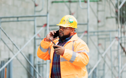 A man in his 30s working at a construction site. He is wearing a hardhat, safety glasses and reflective clothing, holding a walkie-talkie and talking on his mobile phone with a serious expression. He could be a construction worker, foreperson, engineer or building contractor.  A concrete block wall with scaffolding is out of focus behind him. He is mixed race African-American and Pacific Islander.