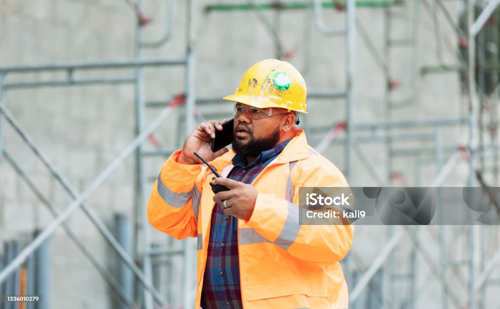 Construction worker at job site talking on mobile phone A man in his 30s working at a construction site. He is wearing a hardhat, safety glasses and reflective clothing, holding a walkie-talkie and talking on his mobile phone with a serious expression. He could be a construction worker, foreperson, engineer or building contractor.  A concrete block wall with scaffolding is out of focus behind him. He is mixed race African-American and Pacific Islander. Construction Worker Stock Photo