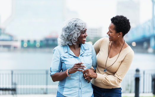 A senior African-American woman in her 60s and her adult daughter, in her 40s, arm in arm outdoors on a city waterfront, a bridge out of focus in the background. They are looking at each other, smiling. The caring daughter is holding her mother's hand.
