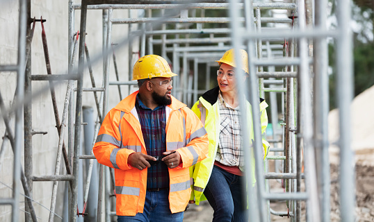 Two multi-ethnic workers at a building construction site, conversing as they walk under scaffolding, wearing hard hats, safety goggles and reflective clothing. The man is mixed race African-American and Pacific Islander, in his 30s. The woman is Hispanic, in her 40s.