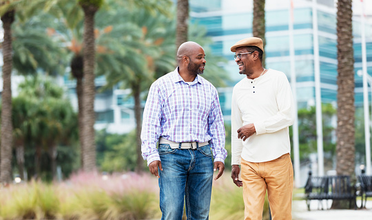 A senior African-American man in his 60s with his son-in-law, in his 40s, walking and conversing in a city park. They are side by side, looking at each other, smiling.