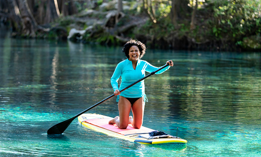 A mixed race Hispanic and Asian woman having fun, kneeling on a stand up paddleboard. She is paddleboarding on a tranquil Florida river with trees covering the riverbank, enjoying nature. She is laughing and looking at the camera.