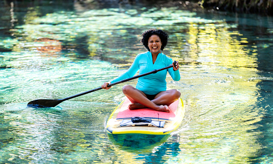 A woman sitting cross-legged on a stand up paddleboard. She is paddleboarding on a tranquil Florida river with trees covering the riverbank, enjoying nature. The mixed race Hispanic and Asian woman, in her 30s, is smiling and looking at the camera.