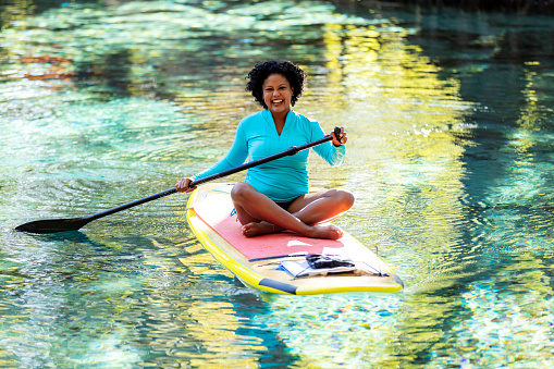 A woman sitting cross-legged on a stand up paddleboard. She is paddleboarding on a tranquil Florida river with trees covering the riverbank, enjoying nature. The mixed race Hispanic and Asian woman, in her 30s, is smiling and looking at the camera.