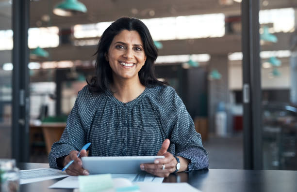 shot of a mature businesswoman using a digital tablet and going through paperwork in a modern office - business woman imagens e fotografias de stock