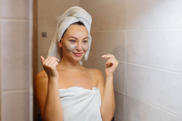 spa i kosmetologia. pielęgnacja skóry twarzy w domu. dziewczyna w łazience nakłada kremową maskę na twarz - clear sky human skin towel spa treatment zdjęcia i obrazy z banku zdjęć