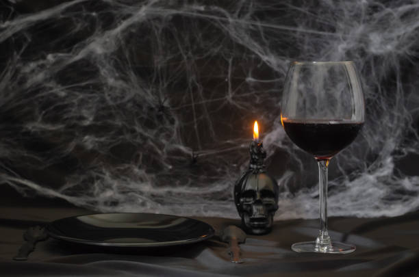 Scary halloween dinner party concept with a glass of red wine Scary halloween dinner party concept with a glass of red wine, black plate, knife, fork and skull candle with cobweb on black background. terrible meal stock pictures, royalty-free photos & images