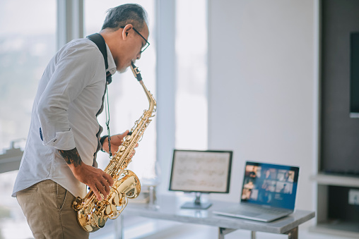 asian chinesse active senior man artist playing saxophone and showing it to his student using laptop at apartment living room in city live streaming