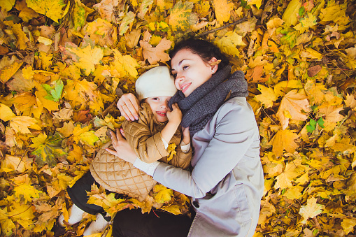 Beautiful young mother and her happy daughter lying on the ground in the forest at autumn. Walks in an autumn park with the kids. Family spending great time playing together with fallen leaves.
