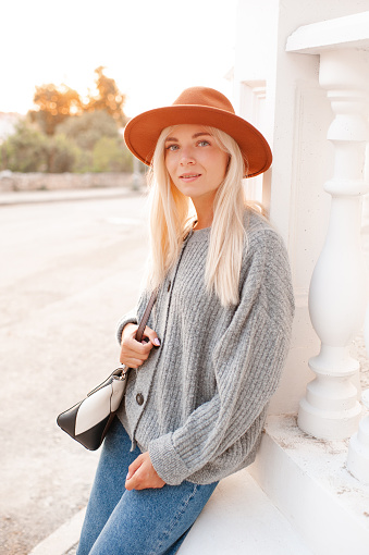 Stylish blonde young adult girl 22-24 year old wear felt hat, denim pants and knit woolen cardigan holding trendy leather bag walk in city street posing over sunset background outdoors close up. 20s.