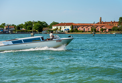 Murano, Italy - June 2, 2021: White water taxi in motion in the Venice lagoon in a sunny spring day in front of the Murano island driven by a man taxi driver, famous for the production of artistic glass. The fastest way to travel in this city. Venice, Italy, Veneto, Europe.