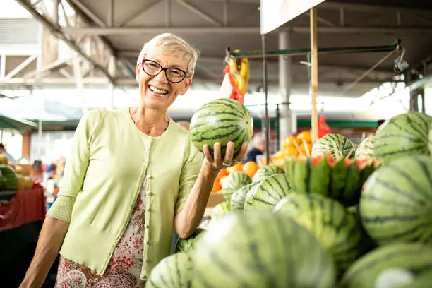 Senior caucasian woman buying fresh organic watermelons and fruit at market place and holding bag full of healthy food.