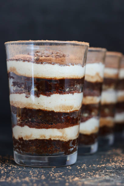 Close-up image diagonal row glasses containing homemade coffee flavoured tiramisu desserts, mascarpone cream, chocolate, sponge cake lady's fingers dipped in espresso coffee layers, dusted with cocoa powder, black background Stock photo showing homemade tiramisu desserts, ready to be eaten. This popular coffee flavour pudding is being served in glasses showing the individual layers of mascarpone cream, chocolate, and sponge cake lady's fingers dipped in espresso coffee. tiramisu glass stock pictures, royalty-free photos & images