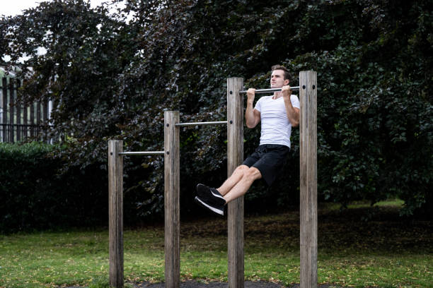 Dedicated young man doing chin-ups at park in rainy weather Three-quarter front view of Caucasian man in white t-shirt, black shorts and shoes, strengthening upper back, arm, and abdominal muscles during weekend workout. Pull-ups stock pictures, royalty-free photos & images