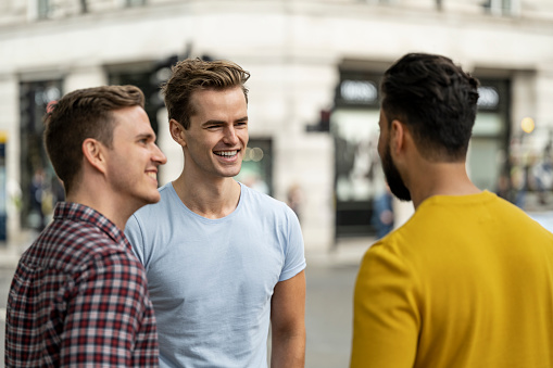 Waist-up view of Caucasian and Indian men in casual weekend attire smiling and talking outdoors in urban environment.