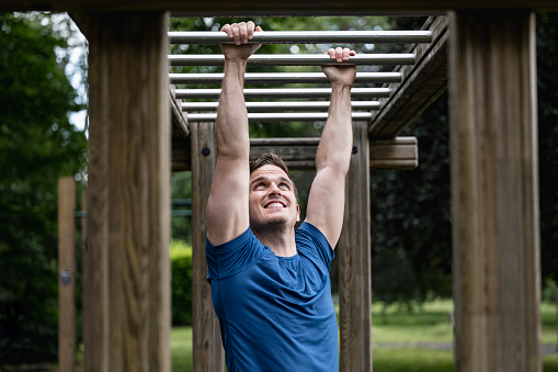 Waist-up front view of man with muscular build wearing blue t-shirt and smiling as he grips alternating bars, improving upper body strength.