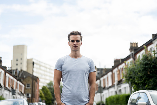 Low angle waist-up view of Caucasian man in light grey t-shirt standing in middle of residential road looking at camera with serious, confident expression.