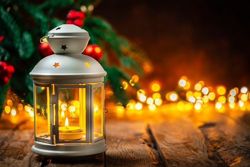 White Christmas lantern with a burning candle shot on rustic wooden table with defocused Christmas tree and lights at background. Selective focus on lantern. The composition is at the left of an horizontal frame leaving useful copy space for text and/or logo at the right.