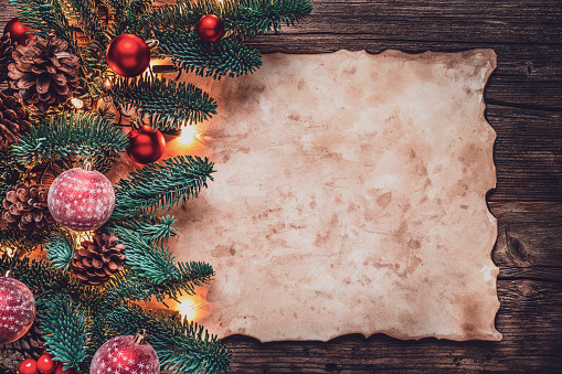 Christmas background with green fir tree branches, Christmas ornaments and lights with a blank parchment on rustic wooden table. The composition is at the left of an horizontal frame leaving useful copy space for text and/or logo.